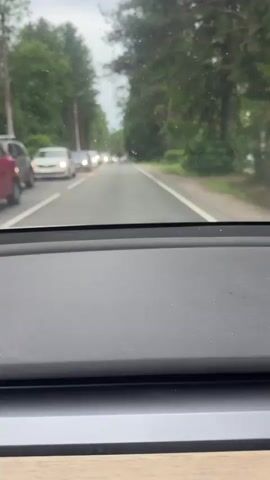This is a first-person perspective video from inside a car driving on a road lined with trees. The road appears to be a two-lane highway with a white line marking the center and the right edge. There is a moderate amount of traffic with cars visible in front, moving in the same direction. The sky is overcast, suggesting cloudy or rainy weather. The trees on either side are lush and green, indicating a temperate climate, possibly summer or late spring. The time of day appears to be daytime, but the overcast sky makes it difficult to determine the exact hour. The dashboard of the car is visible in the foreground, suggesting the video was recorded from the driver's or passenger's seat. There are no people visible outside of the cars in traffic. The overall mood is mundane, depicting a typical drive through a scenic, wooded area with some traffic congestion. No visible text is present in the video. The location context places this scene in Beloostrov, Russia, a town known for its proximity to the Finnish border and its forested landscapes.