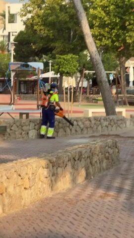 A person wearing a blue cap, yellow safety vest, and blue pants with yellow accents is using a backpack leaf blower to clear debris from a paved walkway. The person is facing left and appears to be moving the leaf blower from right to left. In the background, there is a low stone wall, a tree, and playground equipment. The lighting suggests it is daytime, likely in the late afternoon or early evening due to the warm light and shadows.