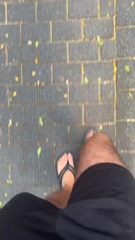 This is a first-person perspective video of someone walking on a paved sidewalk. The scene is outdoors, and the time of day appears to be daytime, with natural light illuminating the path. The ground is made of grey rectangular pavers, scattered with fallen leaves in shades of green and yellow. The individual is wearing dark shorts and black flip-flops, and their bare legs are visible. The movement of the camera suggests the person is walking at a moderate pace.

There are no discernible objects or people other than the person walking. The setting appears to be a public walkway, possibly a park or a street, given the paving and scattered leaves. The weather seems pleasant, as indicated by the attire and the outdoor activity. There are no visible signs or text within the frame. The video captures a simple, everyday moment of someone walking through an outdoor environment. The location context provided, Santa Coloma de Gramenet, Spain, suggests this might be a common walking path in that region.