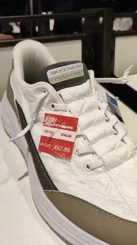 The video shows a close-up of several pairs of shoes on a display shelf in what appears to be a retail store. The first shoe featured is white with a quilted pattern and a brown toe cap. It has white laces and a white sole. A red price tag is attached to the side of the shoe, displaying "RETAIL £55.00" and "OUTLET €52.99". The second shoe visible is white with navy blue accents, including the sole and the inside lining. It also has a red price tag, but the text is too blurry to read. The third shoe is a light brown or tan color with a white sole and blue accents. A red price tag is attached to this shoe as well, with "RETAIL £55.00" and "OUTLET €52.99" clearly visible. The background shows shelves with more shoes and a blue sign with white text, part of which reads "UOMO". The lighting suggests an indoor retail environment.