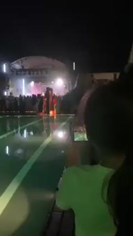 At night, a crowd gathers in front of a stage, reflecting in a pool, as seen from a side perspective where people are using phones to record. The stage is illuminated by bright white lights, and a spotlight shines on the crowd. The pool has a green tint and a white stripe running through its center, adding to the visual spectacle. The atmosphere suggests a live performance or event.