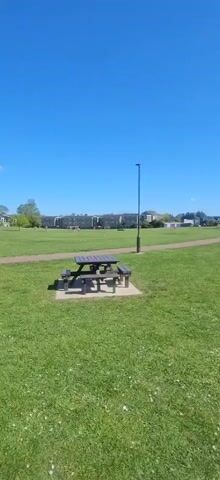 The video presents a panoramic view of a sunny public park in Norwich, United Kingdom. The scene opens with a wooden picnic table with attached benches, set on a small concrete pad, positioned prominently in the foreground on a lush green lawn. A tall, slender pole or lamppost stands just behind the picnic table to the right.

The camera then slowly pans to the left, revealing more of the expansive parkland. A winding dirt or gravel path traverses the mid-ground, separating the immediate foreground from the distant elements. In the background, a cluster of buildings, possibly residential or institutional, with multiple stories, is visible beneath a clear, vibrant blue sky. Numerous trees, some still sparse from early spring, are scattered throughout the park and along the distant tree line. Further to the left in the background, a children's playground with swings and other structures can be faintly discerned.

The weather is bright and clear, indicating a sunny day with very few clouds in the expansive blue sky. No people or specific activities are visible in the park, which appears quiet and serene. The overall impression is one of open space and natural greenery within a developed area. There is no visible text in the scene.