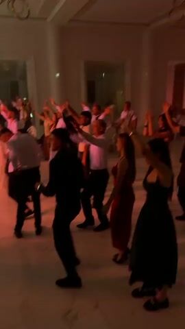 A group of people are dancing in a brightly lit hall. The hall appears to be a banquet hall or event space, with high ceilings and large mirrors. The floor is tiled with a marble-like pattern. In the foreground, a young man in a black shirt and pants is dancing, his arms raised. To his right, a young woman in a green dress is also dancing, her arms raised. Further back, other people are dancing, some in formal attire. There is a band playing in the background, with several musicians playing accordions and other instruments. The lighting changes throughout the video, shifting from bright white to purple and pink. Some of the dancers are holding up what appear to be small lights or phones. In the background, there are tables and chairs, suggesting a catered event.