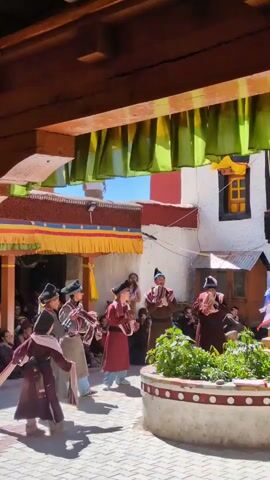 The video shows a courtyard with people in traditional attire, likely monks, performing a ritual dance. There are several individuals in the center of the frame dressed in maroon robes and elaborate hats. They are holding musical instruments and appear to be playing them while moving in a synchronized manner. Some are holding circular objects. The courtyard is paved with interlocking bricks, and there are buildings surrounding it. Colorful banners are hung from the eaves of the buildings, and a small circular planter with green plants is visible in the foreground. Spectators are seated on the ground on the left side of the frame, watching the performance. The sky is blue and the sun is shining, casting shadows on the ground.