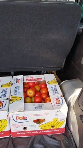 In the back of a vehicle, likely a car or van, two stacked cardboard boxes are visible, filled with produce. The upper box is white with red and blue accents, clearly labeled "Dole Class Extra BANANAS" along with images of bananas. Inside this box, a generous amount of bright red and some yellowish-green tomatoes are packed tightly. The lower box is black with yellow lettering, and the word "yellow" is prominent, also with images of bananas. This box also contains tomatoes, appearing red and green. A dark grey textured surface, possibly the interior of the vehicle's cargo area, occupies the left side of the frame. The lighting suggests it's daytime, with no obvious indications of weather conditions. The overall scene depicts the transport of fresh produce.