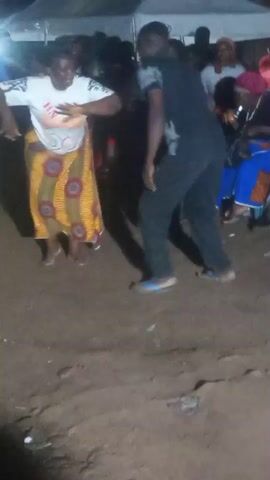 In this video, a group of people are gathered outdoors at night. The ground appears to be dirt or sand. In the foreground, a young child in an orange shirt and red skirt is dancing, with their back to the camera for most of the shot, and then turns to face the camera briefly. To the left, another child in a yellow shirt is partially visible. In the middle ground, a woman wearing a white shirt with graphics and a colorful patterned skirt is dancing. To her right, a man in a black t-shirt and black pants is also dancing. Further back, other people are standing and observing. A white tent structure is visible in the background.