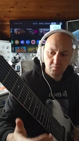 A medium shot depicts a bald, light-skinned man seated, wearing a black hooded sweatshirt with white writing on the chest, playing an eight-string white electric guitar. He’s wearing white headphones connected by a cord to a device not visible in the frame. He appears to be concentrating on his playing, his expression intense. The guitar strap is visible, attached to the guitar body. 

In the background, a large flat-screen TV displays an interface with various app icons and movie posters. The TV sits on a surface behind the man. To the left of the TV is a white cabinet or shelving unit with items displayed on it, including what appears to be a microscope. Behind the chair and to the right of the TV, there is a white surface with assorted small objects. The room has a somewhat cluttered appearance. The ceiling is made of wooden planks.
