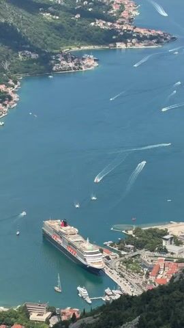 The video offers a high-angle panoramic view of a wide, azure bay surrounded by steep, verdant mountains, likely the Bay of Kotor in Montenegro, consistent with the provided location of Špiljari. Numerous white wakes streak across the water, indicating the movement of many small boats and yachts. A large, multi-deck cruise ship with a red and black funnel is prominently docked near a coastal town, its size dwarfing the surrounding buildings and smaller vessels. Several towns with red-roofed buildings line the bay's edge, nestled between the water and the rising slopes. The sky is partly cloudy, and the overall scene depicts a bustling waterway on a clear day.