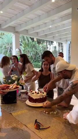 A group of people are gathered around a table outdoors, celebrating a birthday. A red velvet cake with white frosting and berries is being cut by two individuals. One person is holding a knife to cut the cake, while another person guides their hand. Other people stand around the table, some holding drinks and conversing. A balloon with Hebrew writing is visible in the background. The setting appears to be a patio with palm trees and lush greenery. The lighting suggests it is daytime.