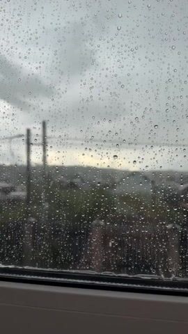 This is a short, vertical video clip showing rain hitting a window. Through the window, a blurred outdoor scene is visible, including houses, trees, and utility poles against a cloudy sky. The focus is on the raindrops clinging to and running down the glass.