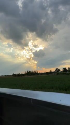 A scenic view of a cloudy sky with sunbeams piercing through the clouds. The sun is low in the sky, casting a warm glow. In the foreground, there is a green field with trees on the horizon. The video appears to be taken from a moving vehicle, as indicated by the blur of the foreground.