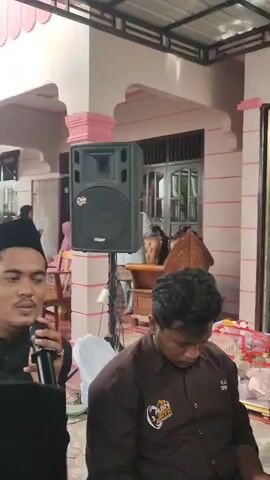 The video shows a group of young men gathered indoors, possibly for a cultural or religious event. In the foreground, a man wearing a brown shirt with a logo is seated, looking down. Behind him, two other men in traditional attire, including black caps and patterned shirts, are holding microphones and appear to be singing or speaking. A third man in similar attire is visible on the right, also holding a microphone. A sound mixing board is prominently placed in the lower right corner, with wires and equipment suggesting a live audio setup. A basketball hoop is mounted on the wall in the background, adding an unexpected element to the scene. The environment appears to be a covered outdoor space or a room with large sliding doors. The lighting suggests it is daytime.