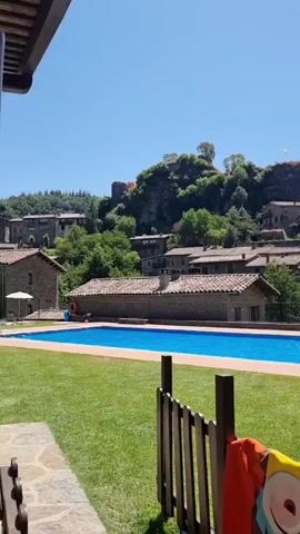 The video shows a sunny day with a clear blue sky. In the foreground, a short wooden fence with some colorful towels draped over it is visible. Beyond the fence is a well-maintained green lawn. Further back, a bright blue swimming pool is the main focus. Several people are in or around the pool, including children playing and adults relaxing. Some are sitting on the edge of the pool, while others are in the water. In the background, there is a cluster of old stone buildings with terracotta roofs, typical of a European village. These buildings are situated on a hillside. At the very top of the hill, a rocky outcrop with some structures, possibly ruins or a castle, is visible. The overall scene depicts a peaceful and idyllic setting, likely a resort or a holiday home, enjoying a warm summer day.