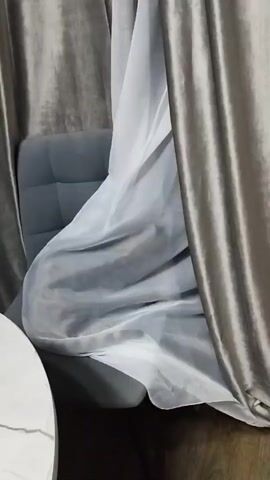 A gray tabby cat is visible through a sheer white curtain, lying on a gray upholstered chair. The cat is mostly hidden by the curtain and the chair. Two heavier gray curtains frame the scene on either side. A portion of a round, light-colored table with a marble-like pattern is visible in the lower left corner. The lighting is dim.