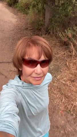 A woman with short brown hair and sunglasses is walking on a dirt path in a wooded area. She is wearing a light blue hooded shirt. The path is surrounded by dry grass and trees. The camera appears to be held by the woman, creating a first-person perspective.