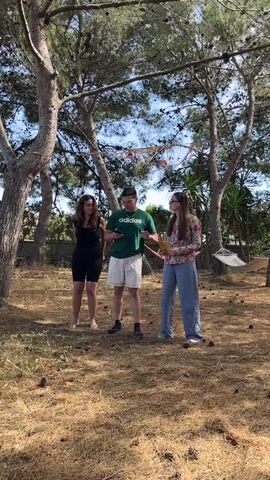 Three people, two women and one man, are standing in a yard that is covered with pine straw and pine cones. Several tall trees surround the yard, with a hammock visible hanging from two of the trees in the background. The man is wearing a green t-shirt, white shorts, and sneakers. He is looking down at his phone. One of the women is wearing a black tank top and black shorts, and is barefoot. The other woman is wearing a light-colored floral shirt and loose-fitting blue pants. She is holding a paper fan in her hand. The weather appears to be sunny and bright. The man turns to the woman on the right and gestures with his left arm. The woman on the left also raises her left arm.