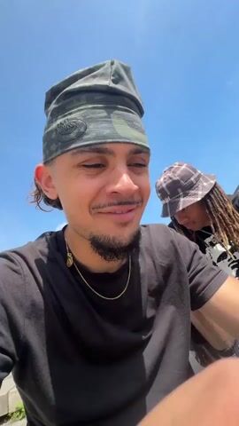 This video captures a skateboarding sequence in Lyon, France, featuring two men under a clear blue sky.

The opening shot is a close-up of a man with a goatee, mustache, and short dark hair styled in a mullet. He wears a camouflage head covering, a black t-shirt, and a gold chain with a medallion, smiling subtly while looking to his right. Partially visible behind him is another person with dreadlocks and a plaid bucket hat.

The subsequent clips show a second man performing a skateboard trick. He wears a black "ACDC" sleeveless t-shirt, dark baggy pants, blue and white sneakers, sunglasses, and a dark baseball cap. He is captured skateboarding down a flight of concrete stairs, extending his arms for balance as he descends. A low-angle shot shows him landing the trick cleanly. The setting includes modern concrete buildings, one with a prominent glass roof structure. On a nearby wall, the text "BIBLIOTHÈQUE" and "DINER" is visible. The final shot shows the skateboarder moving away from the stairs on a paved surface, appearing to kick up the back of his board. The entire sequence takes place in bright daylight, indicating clear and sunny weather.
