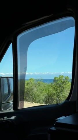The view from inside a vehicle shows a window looking out to a body of water, a tree-lined shore, and snow-capped mountains in the distance under a blue sky.  The vehicle's side mirror is visible in the left portion of the window.