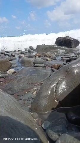 The video captures a close-up view of waves crashing on a rocky shore. The camera is positioned low, near the water's edge, focusing on the wet, dark rocks and the foamy white water as it recedes. In the background, larger waves are visible against a bright blue sky with scattered clouds. The Infinix NOTE 40Pro+ watermark is present in the bottom left corner.