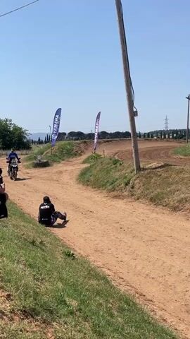 The video shows a dirt track on a sunny day. In the foreground, a person wearing a black jacket and black pants is sitting on the ground, seemingly taking a break. To the left, several people are watching. The dirt track curves into the distance, flanked by grassy areas and two blue Yamaha flags. Several motorcyclists, wearing helmets and riding gear, are approaching on the track. In the background, there's a rural landscape with trees and a power line. The scene is framed by an electric pole.