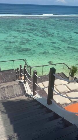 The video shows a view from the top of a set of wooden stairs leading down to a wooden deck overlooking a clear, turquoise ocean. The stairs have a wooden railing supported by white rope. The deck has clear glass railings on either side and several white lounge cushions arranged on the surface. In the ocean, there are patches of dark spots, possibly coral or rocks. In the distance, the ocean horizon is visible under a clear blue sky. A person is visible swimming in the ocean.
