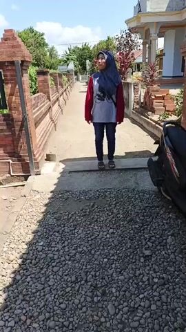 A young woman wearing a blue hijab and a red and gray shirt stands on a concrete path. She is facing forward and looking up. To her left is a brick wall and to her right is a building. In the foreground, the path is covered in gravel. A motorcycle is parked to the right of the path. The sky is blue and the sun is shining.