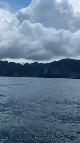 The video shows a body of water with a distant mountain range under a cloudy sky. The water appears choppy and reflects some sunlight. The mountains are dark and appear to be heavily forested or rocky. The clouds are large and white, with some darker areas suggesting potential rain. The camera appears to be moving, as indicated by the slight shift in perspective and the gentle movement of the water. There are no people or identifiable objects in the foreground or midground.