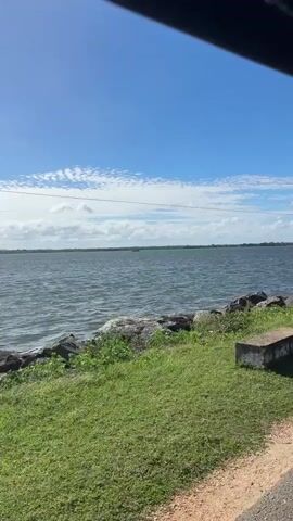 A vehicle is traveling down a paved road beside a large body of water, possibly a lake or a reservoir. The water is choppy, with small waves visible. The shoreline is lined with large rocks and grassy vegetation. The sky is blue with scattered white clouds. A single bird is flying in the distance.