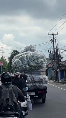 The video shows a truck overloaded with large, puffy items that appear to be mattresses or bedding, wrapped in clear plastic with a dark floral pattern. The cargo is secured with ropes and netting. The truck is driving on a street with other vehicles, including motorcycles and cars. Power lines and buildings line the street, suggesting an urban or suburban environment. The sky is cloudy. The license plate on the truck reads "DK 8352 FZ". The camera angle is from behind and slightly to the side of the truck, possibly from another moving vehicle.