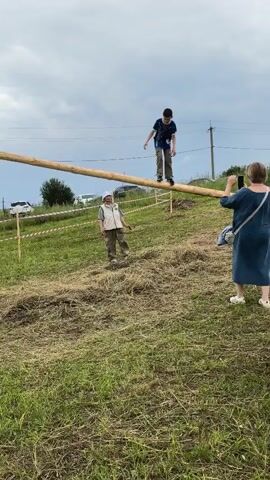 A young boy wearing a dark blue t-shirt and khaki pants attempts to walk across a long, thick wooden pole suspended horizontally above a grassy slope. He loses his balance and jumps off the pole. Another person, possibly an adult, wearing a light-colored vest and khaki pants, stands on the slope below the pole, watching. To the right, a woman in a dark blue dress is standing and holding a phone, seemingly recording the event. The environment is an outdoor grassy area with a cloudy sky.
