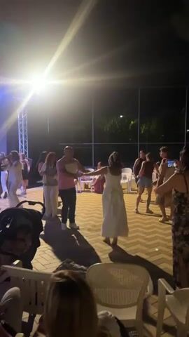 The video shows a group of people dancing outdoors at night. A bright spotlight illuminates the dancing area, creating long shadows on the patterned brick ground. Several individuals, including men and women of various ages, are engaged in dancing, some in pairs and others in a group. One woman in a white dress is prominently featured dancing with a man in a pink shirt. In the background, there are chairs and a fence, with trees and some lighting visible beyond the fence. A stroller is parked on the left side of the frame. The overall atmosphere appears to be festive and social, likely at an event or party.