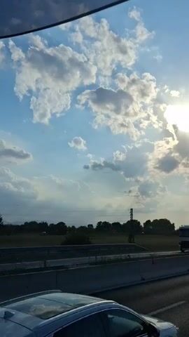The video begins with a shot of a white truck and a dark colored car on a highway with a bright sun partially obscured by clouds in the background. The camera then pans to the right, showing more cars on the highway, including a white SUV in the foreground, and the sun and clouds in the sky. The video continues to show the highway with traffic and the sky. The final shot is of the highway with cars and the sky.