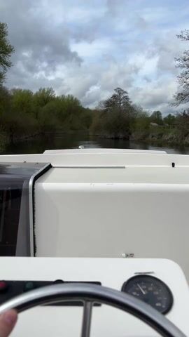 The video shows a first-person point-of-view shot from the helm of a white boat as it navigates a river. The boat's steering wheel and a dashboard with gauges are visible in the foreground. A hand is lightly placed on the steering wheel. The river is surrounded by trees on both banks, and the sky is overcast with gray clouds. The water is calm and reflects the sky and trees. The boat is moving forward at a slow pace, as indicated by the speedometer reading.