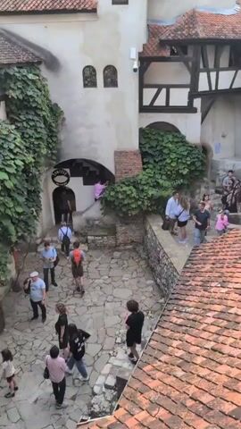 The video shows an aerial view of a medieval castle. People are walking around in the courtyard and on the stairs. The castle has a tall, conical tower and a tiled roof. There is greenery growing on the walls of the castle. The sky is cloudy.