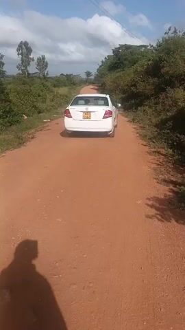 The video shows a white car driving down a dirt road. The road is surrounded by trees and bushes. The sky is blue with white clouds. The camera is positioned behind the car, looking down the road. The shadow of a person holding the camera is visible in the bottom left corner of the frame. The audio contains a person speaking a language that is not English.