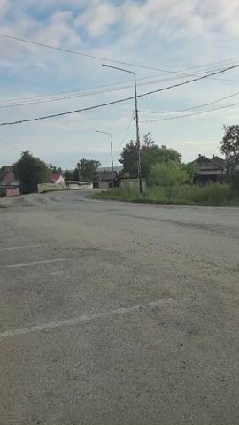 The video shows a rural road with a light gray, gravel surface. A white SUV is driving away from the camera on the right side of the road. The car is in motion. The environment is open with some trees and buildings in the background. The sky is partly cloudy. Power lines are visible overhead.