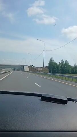 The video shows a view from the dashboard of a car driving on a highway. The highway is paved with asphalt and has white lane markings. There are guardrails on the sides of the highway. In the distance, there is an overpass. The sky is blue with some clouds. There are streetlights along the highway. There is a black object on the dashboard. The car appears to be moving at a moderate speed. The overall environment is a typical highway landscape.