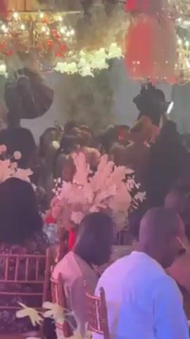 The video appears to be taken at a festive event, possibly a wedding or large party. People are milling about, many of them moving to the music playing in the background. Some are wearing formal attire. The area is decorated with chandeliers and flower arrangements. A waiter in a black uniform is carrying a tray of drinks through the crowd. Blue stage lighting adds to the atmosphere.
