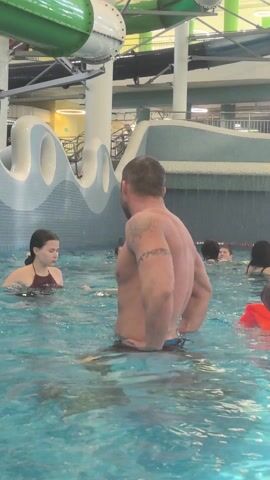 The video shows a man standing in a swimming pool. He is facing away from the camera, and his back is visible. He has short, graying hair and a tattoo on his left arm. There are several other people in the pool, including women and children. The pool is part of an indoor water park, with water slides and other attractions visible in the background. The water is a light blue color.