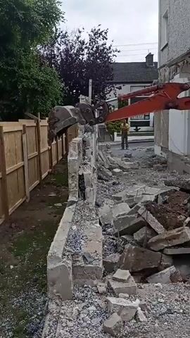 An excavator with an orange boom and a bucket is demolishing a concrete wall. The excavator's bucket is actively breaking apart the wall, causing concrete debris to fall onto the ground. The wall appears to be a long structure, with sections already reduced to rubble. To the left of the wall is a wooden fence and a patch of grass. To the right of the wall, the side of a building is visible. In the background, a person wearing a yellow vest is standing near the demolition site, observing the work. The sky is overcast.