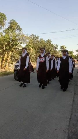 A group of young girls in uniform are lined up and appear to be walking or marching. They are wearing brown dresses with white shirts and brown headscarves. Some have bibs with numbers. The scene appears to be outdoors on a paved surface, possibly a street or path, with trees and vegetation in the background. The lighting suggests it is daytime. One girl in the foreground has a bib with the number 076.