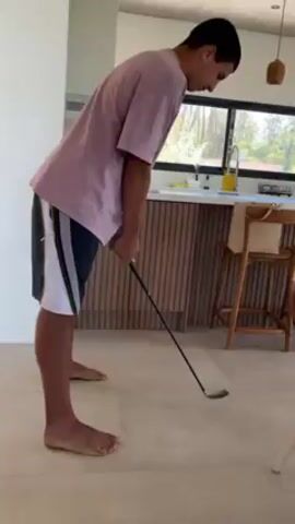 A young man stands barefoot on a tiled floor in a bright, modern interior and practices a golf swing. He is wearing a pale pink t-shirt and dark shorts with white stripes down the sides. He bends forward, gripping a golf club, then swings it up behind his head. He turns to face the camera, saying something, then walks away. Behind him is a kitchen area with white countertops, dark cabinets with vertical slats, a sink, and a yellow pitcher. A wooden stool with a cushion is beside him. A window lets in natural light. A pendant lamp hangs from the ceiling.