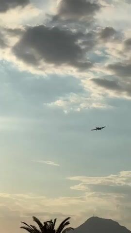 The video shows a small propeller plane flying in the sky. The sun is visible and shining brightly, with lens flares. There are clouds in the sky, some of which are dark and appear to be stormy. In the foreground, there is a palm tree and a light pole. The plane appears to be in descent, possibly approaching an airport. The scene appears to be taking place at sunset or sunrise due to the lighting conditions.