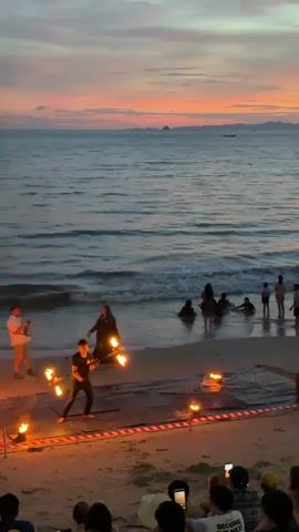 A fire dancer performs on a sandy beach as the sun sets over the ocean. The dancer is wearing a dark outfit and is twirling two flaming objects, creating a circular pattern of light. Several spectators are gathered on the beach to watch the performance, some are sitting and others are standing. Some people are in the shallow water near the shore. The sky is filled with soft colors of orange, pink, and purple as the sun dips below the horizon. The waves gently roll onto the shore, and a small boat is visible in the distance. Red and white tape marks the perimeter of the performance area.