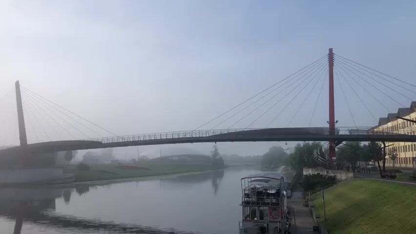 A wide shot shows a modern cable-stayed bridge spanning a calm river on a foggy day. The bridge has two red pylons, one on each side of the river, supporting the roadway with cables. A walkway is visible on the bridge. The river reflects the overcast sky and the bridge. On the right bank, there is a grassy area with trees and a building. On the left bank, there is a building with a curved roof structure, and a grassy area with some colorful designs. A small boat or barge is docked on the right bank in the foreground. The fog obscures the background, creating a hazy atmosphere. No people are visible in the video.