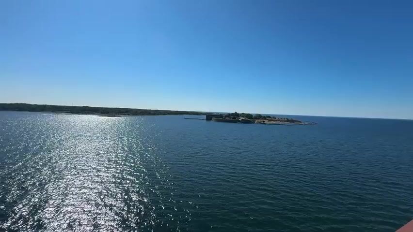 The video is a wide shot of an island in the middle of a large body of water. The island has a fort or castle-like structure on it. The water is choppy with sunlight reflecting off of it. The sky is clear and blue. The camera appears to be moving slightly, possibly from a boat. There are no people visible in the video.