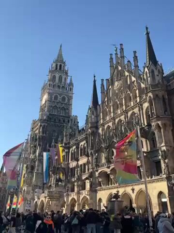 The video shows the Neues Rathaus, or New Town Hall, in Munich, Germany. The building is a large, ornate Gothic Revival structure with a tall clock tower. Colorful flags are hanging in front of the building.  A crowd of people are gathered in the square in front of the Rathaus. The sky is clear and blue.
