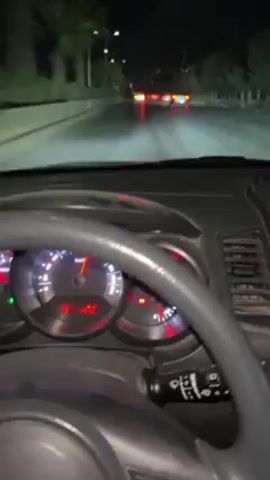 The video shows a car's dashboard at night. The speedometer indicates the car is moving, and other dashboard lights are illuminated. Through the windshield, one can see a dark road with other vehicles ahead, their tail lights visible. Streetlights are present along the roadside. The perspective is from the driver's seat.