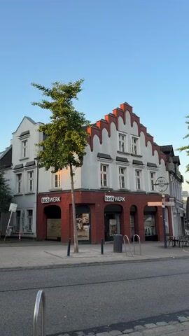 The video shows a street view in a European town during what appears to be early morning or late afternoon due to the low angle of the sun. The camera pans slowly from left to right, capturing the facade of a brick and white building with distinctive stepped gables. The building has a "back Werk" bakery on the ground floor. A young tree stands in front of the building. Further down the street, more buildings are visible, some with awnings and outdoor seating. A white van is parked on the road. A lamppost with a hanging flower basket is also prominent. The sky is clear blue. No people are visible in the video.