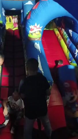 Several children are playing on a large inflatable slide inside what seems to be an indoor play area. The slide is decorated with images reminiscent of the Super Mario universe, including mushrooms and Mario himself. The slide's surface is red, and the surrounding inflatable structure is primarily blue. The children are ascending the stairs to the slide and sliding down. One child is wearing a black shirt and grey pants. There appears to be an adult or older child standing near the slide, possibly supervising. The lighting in the area seems dim, and there's noticeable movement throughout the video.